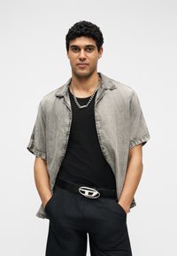 Νέος άνδρας με σγουρά μαλλιά που φοράει γκρι denim πουκάμισο με κοντά μανίκια πάνω από μαύρο μπλουζάκι, μαύρο παντελόνι, αλυσίδα στο λαιμό και ζώνη με μεγάλο ασημένιο αγκράφα.