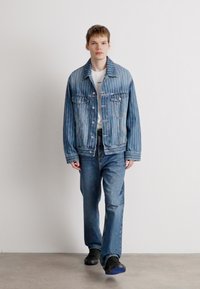 Τζάκετ από τζιν με κάθετες ρίγες, μπροστινό κλείσιμο με κουμπιά, δύο θηκών στο στήθος; σε συνδυασμό με ελαφρύ μπλουζάκι, τζιν παντελόνι με ίσια γραμμή και αθλητικά παπούτσια.