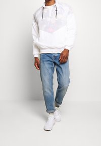 Felpa bianca con mezza cerniera e motivo floreale sottostante, abbinata a jeans azzurri e scarpe da ginnastica bianche. Cordini neri visibili.