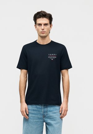 Mann trägt ein schwarzes Tommy Hilfiger T-Shirt mit Logo auf der Brust und hellblaue Jeans, steht vor einem schlichten hellen Hintergrund.
