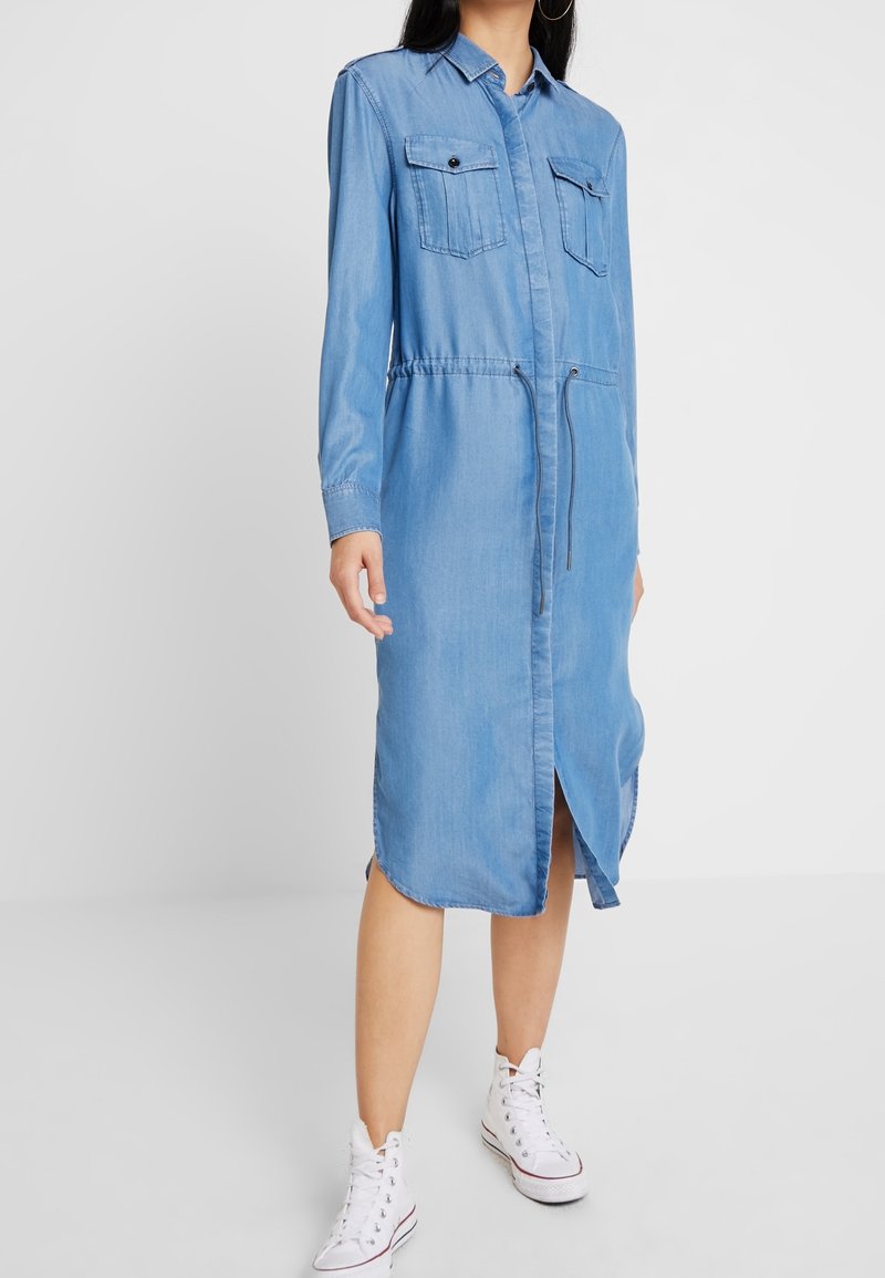 Robe en denim bleu clair à manches longues, deux poches poitrine et taille à cordon. Longueur genou avec une fente avant. Portée avec des baskets blanches.