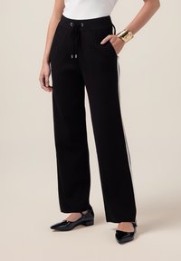 Pantalons larges noirs avec une taille élastique à cordon, poches latérales et rayures blanches sur les côtés ; associés à des chaussures noires brillantes.