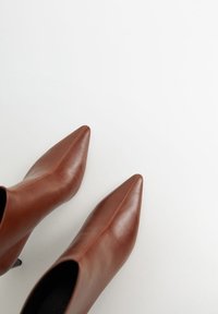 Bottes basses en cuir marron avec bouts pointus, texture lisse, coutures minimales et talons fins sur fond blanc.