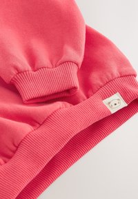 Koralliroosa pusa, millel on soonikkoes mansetid ja alaäär, pehme tekstuur ning küljes väike karu logo etikett.