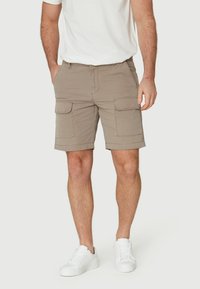 Pantalones cortos beige de cargo hechos de algodón, con grandes bolsillos laterales, bolsillos frontales y detalles de costura; usados con deportivas blancas.