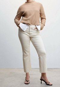 Suéter beige de manga larga superpuesto sobre una camisa blanca, combinado con jeans rectos crudos y tacones negros con tiras. Textura suave.
