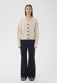 Femme portant un cardigan beige boutonné, un pantalon large bleu marine et des sandales noires, debout devant un fond blanc uni.
