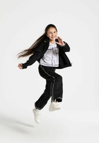 Czarna kurtka zapinana na zamek noszona na białej koszulce z grafiką i czarnych dresach z białymi akcentami, w połączeniu z białymi trampkami. Włosy są długie i swobodne.