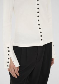 Biała bluzka z długim rękawem z pionowymi czarnymi guzikami z przodu i na mankietach, połączona z czarnymi spodniami o wysokim stanie z plisami.