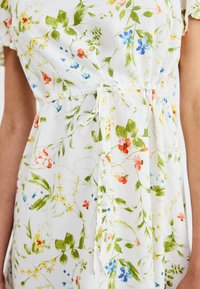Robe fleurie en tissu blanc ; ornée de feuilles vertes et de fleurs multicolores, cintrée à la taille avec un lien, manches courtes.