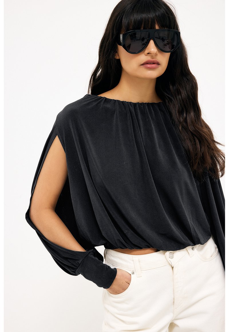 Femme portant de grandes lunettes de soleil noires et un haut noir ample à manches longues avec des découpes sur les manches, associé à un pantalon blanc.