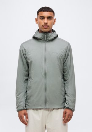 Hombre con chaqueta con capucha gris clara con cremallera y logo de Salomon en el pecho, de pie frente a un fondo blanco liso.