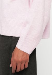 Ανοιχτού ροζ πουλόβερ κατασκευασμένο από μαλακό ύφασμα, με ριγωτές μανσέτες και τελείωμα, συνδυασμένο με σκούρο γκρι παντελόνι, που παρουσιάζει μια λεία, υφασμάτινη υφή.