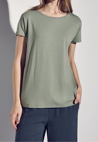 Kortärmad olivgrön t-shirt gjord av mjukt material; har rund hals och avslappnad passform, i kombination med mörka, löst sittande byxor.