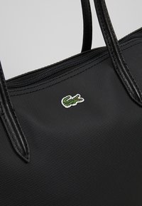 Sac à main en nylon noir avec une surface texturée, doté d'une fermeture zippée sur le dessus et de deux poignées. Logo de crocodile vert brodé affiché de manière proéminente.
