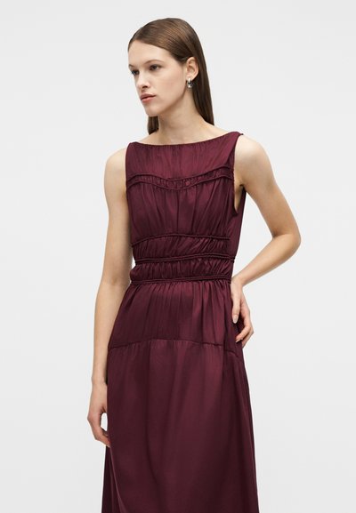 Νεαρή γυναίκα με ίσια καστανή μαλλιά, ντυμένη με αμάνικο σκουρομωβ φόρεμα με ρυτίδες, στέκεται με το ένα χέρι στη μέση μπροστά σε απλό φωτεινό φόντο.