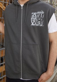Hombre vestido con una sudadera sin mangas de color gris oscuro con cremallera y un gráfico de "Aceleración Sin Parar" y diseño de objetivo en el pecho, de pie junto a una valla de metal.