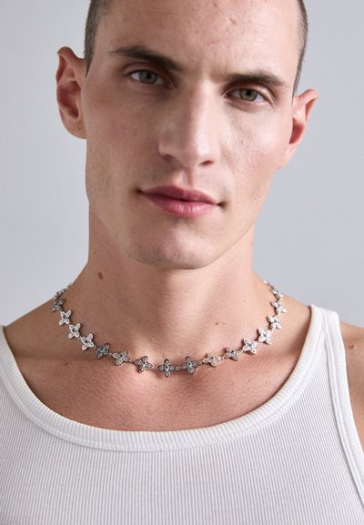 Young man wearing a silver necklace with floral-shaped links and a white ribbed tank top against a plain background.