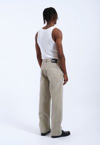 Beige Denim-Jeans mit einem lockeren Schnitt, Fünf-Taschen-Design und schwarzem Label am Bund. Trägt ein weißes Tanktop, von hinten betrachtet.