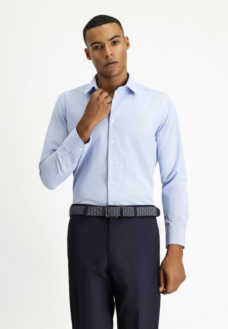 Camicia da uomo a maniche lunghe di colore azzurro chiaro, con un motivo a strisce sottili. Presenta un colletto punta e bottoni ai polsini, abbinata a pantaloni scuri e una cintura intrecciata.