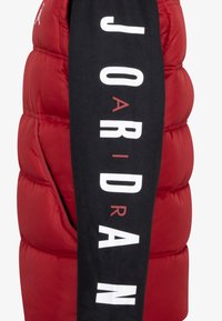 Rød polstret vest med en sort ærme, med fed hvid skrift "JOARIDAN" og et lille logo. Glat tekstur med et voluminøst design.