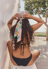 Zwarte bikini onderkant met een strikontwerp aan de achterkant. Haar vastgebonden met een bloemige roze sjaal met groene bladeren en rode bloemen. Gestructureerde muur en stenen pad zichtbaar.