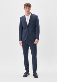 Tummansininen puku, jossa on teksturoitu kangas, yksirivinen bleiseri ja slim fit -housut, yhdistetty valkoiseen t-paitaan ja mustiin loafer-kenkiin.