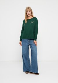 Sudadera verde con cuello redondo y logo bordado de "Gap", combinada con jeans de pierna ancha en azul y zapatos negros tipo slip-on.