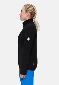 Black fleece jacket with high collar, zip pockets, and logo on the left sleeve. Smooth texture, fitted design, worn with blue pants.