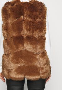 Gilet in ecopelle marrone con design a patchwork, caratterizzato da una consistenza morbida e pelo di lunghezze variabili. Indossato sopra una maglietta bianca a maniche lunghe.