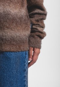 Maglione di lana marrone con una texture morbida e soffice e polsini a coste, abbinato a jeans in denim blu. Etichetta visibile sul maglione.