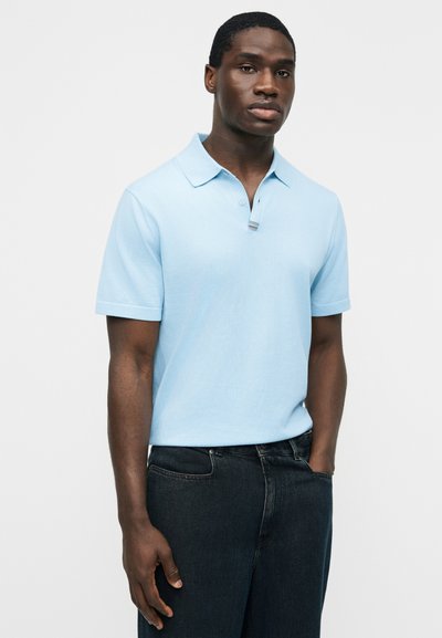 Jeune homme portant un polo bleu clair à manches courtes et un jean noir, debout avec une main dans la poche devant un fond uni.
