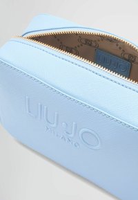 Lyseblå syntetisk taske med en tekstureret overflade, der har en guld lynlås og prægede logo. Indvendigt har den et mønstret for.