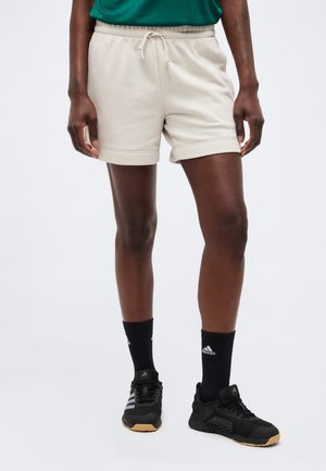 Persoon draagt beige Adidas-short met trekkoord, zwarte Adidas-sokken en zwarte sportschoenen, staand tegen een effen achtergrond.