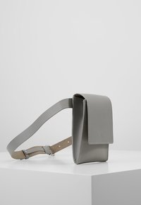 Šedá kožená crossbody taška s klopou, nastavitelným popruhem a hladkým povrchem. Minimalistický design s čistými liniemi a bez viditelného kování.