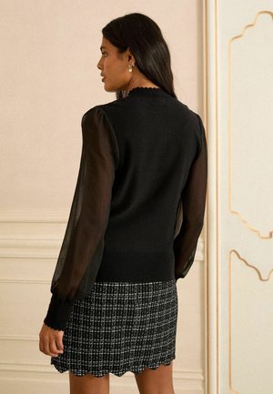 Černý pletený svetr s průhlednými nafouknutými rukávy, se zvlněným výstřihem. Spárováno s černobílou kostkovanou sukní se zvlněným lemem.