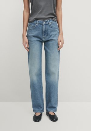 Osoba ubrana w jasnoniebieskie jeansy o prostych nogawkach, ciemnoszary bezrękawnik i czarne płaskie buty, stojąca na jednolitym jasnym tle.