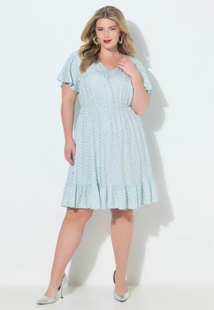 Käyrä nainen vaaleansinisessä kukallisessa polvipituissa mekossa, jossa liehuvat hihat ja hopeanväriset korkokengät, poseeraa valkoista taustaa vasten.