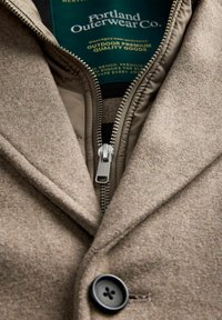 Lysebrun frakke i uldblanding med lynlåslukning, inderfor i beige, sort knapdetalje og label med teksten "Portland Outerwear Co."