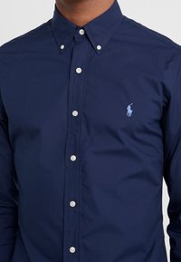 Marinblå skjorta med knappar, tillverkad av mjukt bomullstyg; har en klassisk krage, vita knappar och en ljusblå broderad logotyp på bröstet.