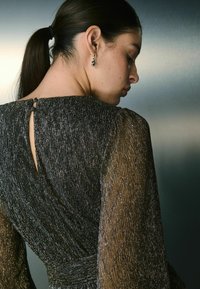 Schwarzes und silbernes metallic Kleid mit transparenten Puffärmeln, rückseitigem Schlüsselaugen-Detail und strukturiertem Finish, von hinten mit unscharfem Hintergrund gezeigt.