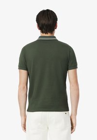 Ciemnozielona koszulka polo z krótkimi rękawami, żebrowanym kołnierzykiem z białymi paskami, gładką fakturą i małym logo blisko dołu.