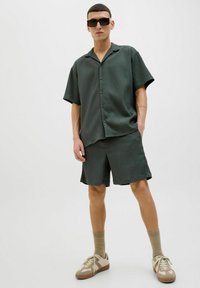 Chemise à manches courtes à rayures fines vert foncé et short assorti, stylisés avec des baskets beige et des chaussettes côtelées beiges. Lunettes de soleil carrées foncées portées.