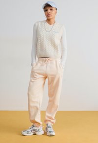 Chaleco de punto beige claro con patrón de trenzas, camisa blanca de manga larga, joggers melocotón y zapatillas grises con detalles en rosa. Gorra azul.
