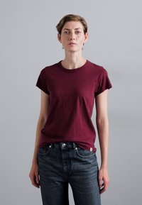 Bordeauxfarbenes Baumwoll-T-Shirt mit kurzen Ärmeln und rundem Halsausschnitt, kombiniert mit dunklen Jeans. Schlichtes Design, weicher Stoff und klassischer Schnitt.