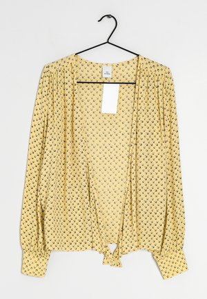 Blouse jaune à manches longues avec un petit motif floral noir, manches froncées et nœud à l'avant, suspendue sur un cintre noir sur fond blanc.