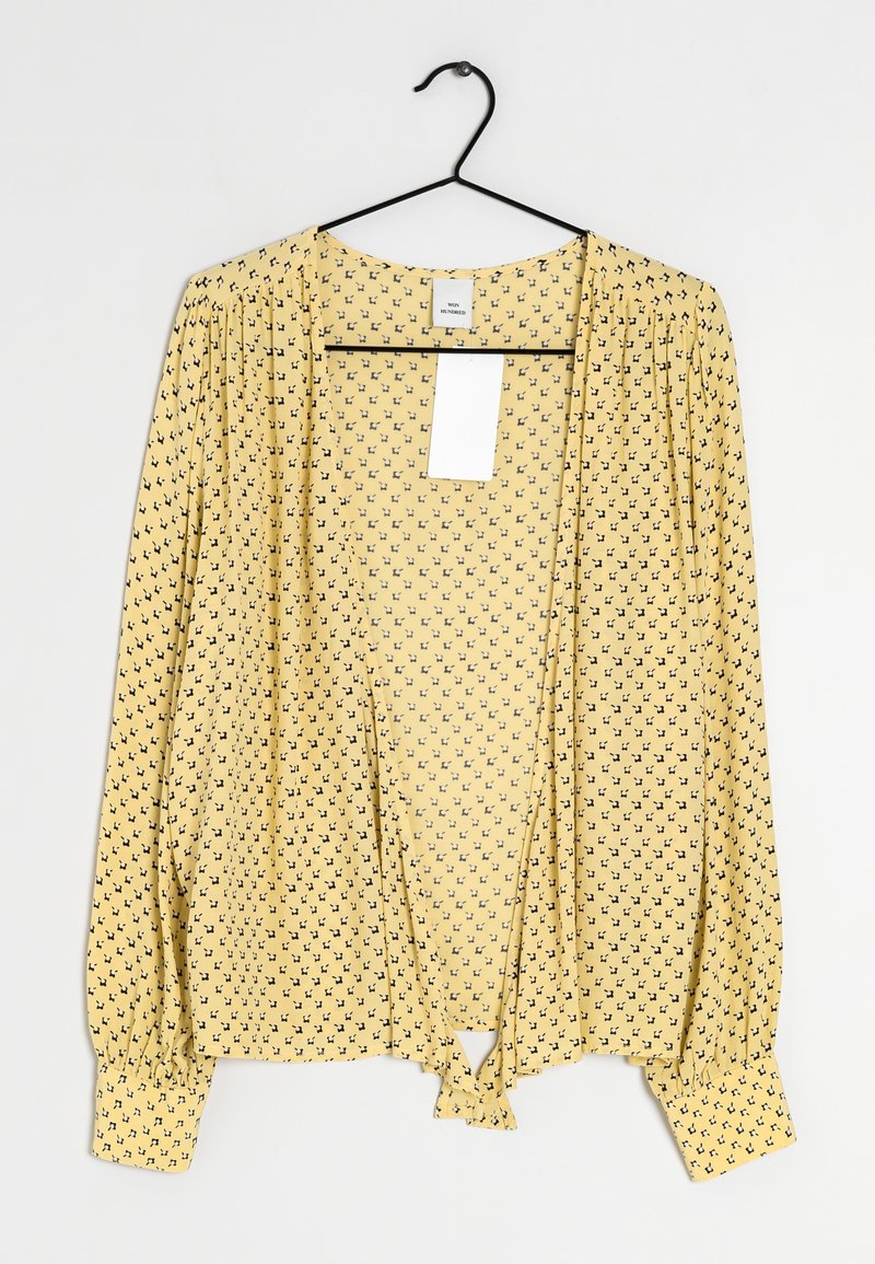Blouse jaune à manches longues avec un petit motif floral noir, manches froncées et nœud à l'avant, suspendue sur un cintre noir sur fond blanc.