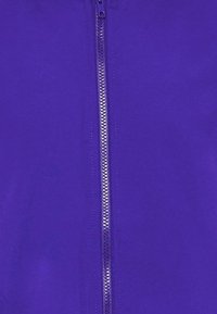 Lilla zip-up jakke med en glat, skinnende tekstur og en central lynlås. Stofdet ser letvægts ud og har ingen synlige mønstre.