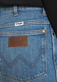 Jeans di denim blu con una toppa in pelle marrone con la scritta "Wrangler" in rilievo e un motivo di cucitura a zigzag sulla tasca posteriore.