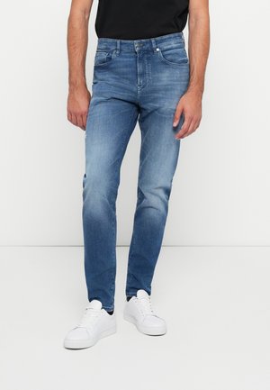 Man wearing slim-fit blue jeans and white sneakers, standing against a plain white background with arms relaxed at sides.